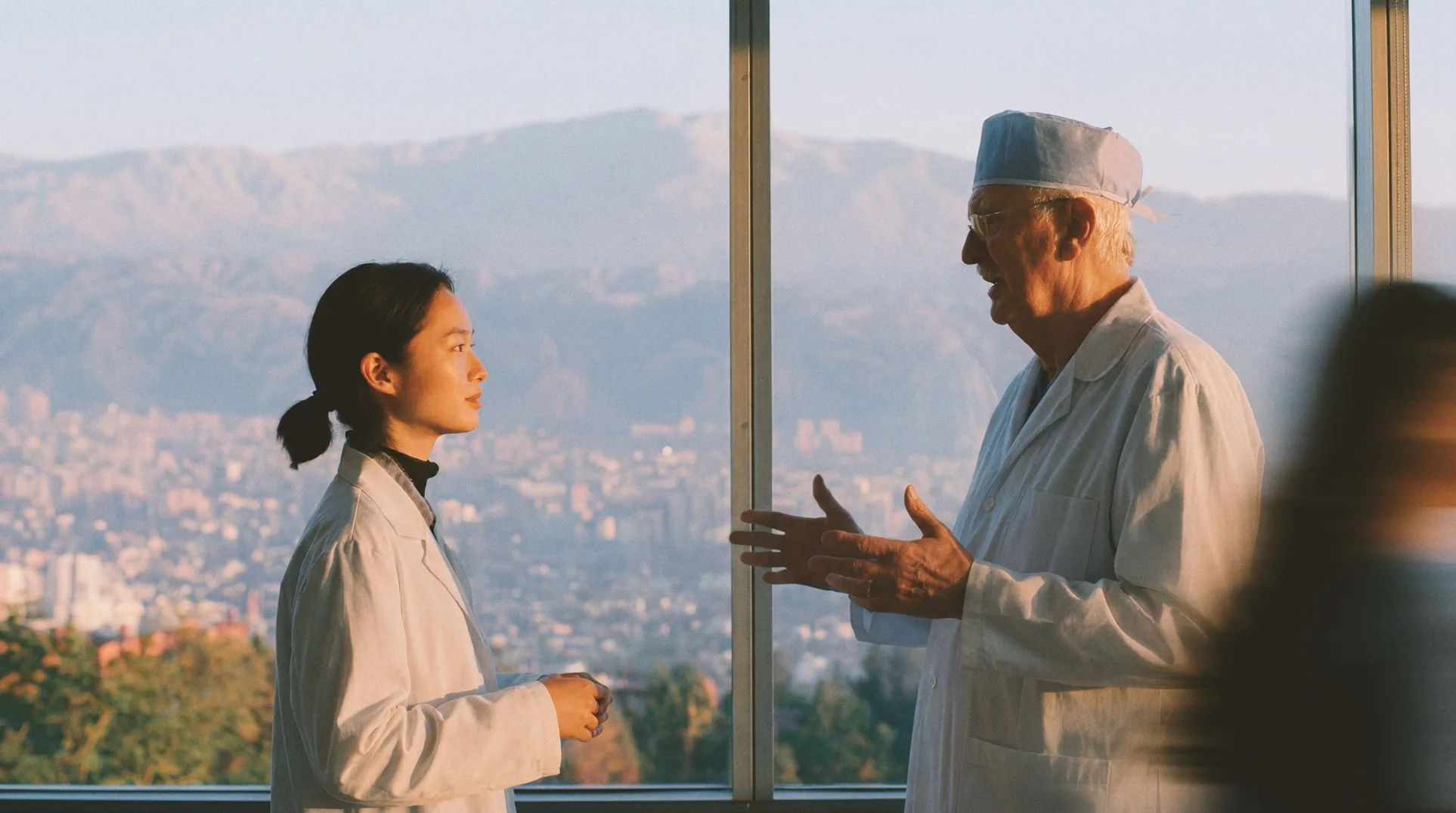 Two clinicians in white coats talking by a window at dusk, a city behind them